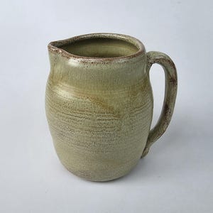 May include: A ceramic pitcher with a rounded body and a single handle. The surface features a mottled light green and beige glaze with visible horizontal texture lines and a darker brown rim and handle edge, creating a rustic, earthy appearance.