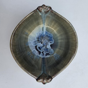 May include: A ceramic bowl with a pointed, leaf-like shape and two decorative handles. The interior features a glossy glaze in shades of olive green and brown, with a prominent, star-like crystalline pattern in deep blue and white at the center.