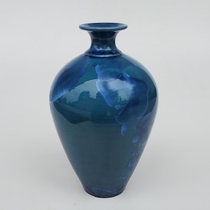 May include: A tall, glossy ceramic vase in shades of blue. The vase has a rounded body that tapers to a narrow base and a flared rim. The surface features a mottled pattern of lighter and darker blues.