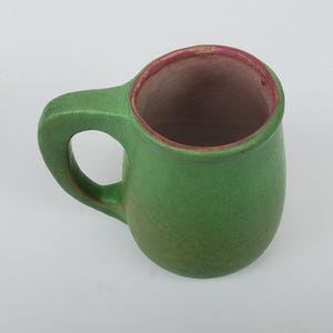 May include: A handmade green ceramic mug with a rounded body and a sturdy handle. The interior is a contrasting brown with a red rim. The mug is designed for hot beverages and is suitable for everyday use.