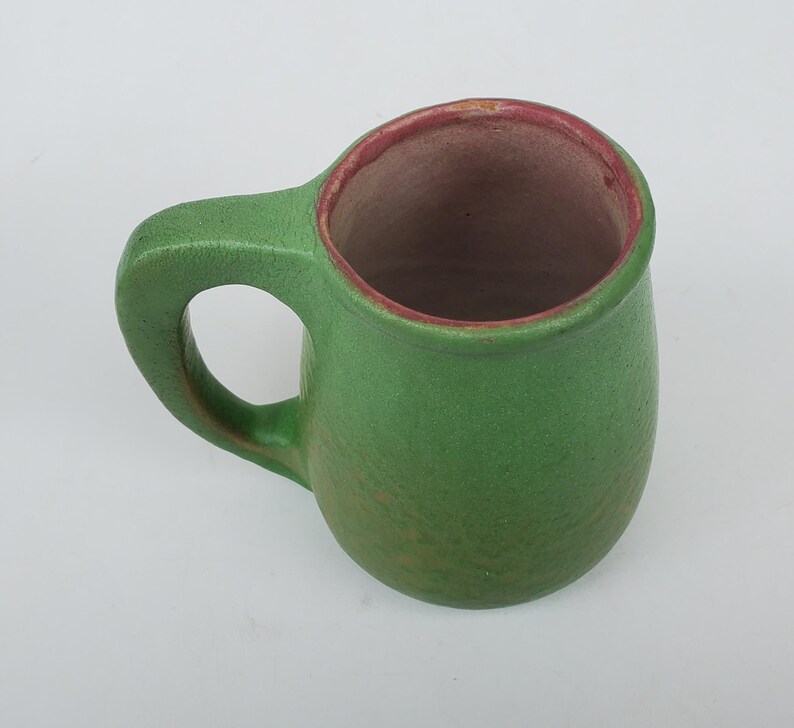 May include: A handmade green ceramic mug with a rounded body and a sturdy handle. The interior is a contrasting brown with a red rim. The mug is designed for hot beverages and is suitable for everyday use.