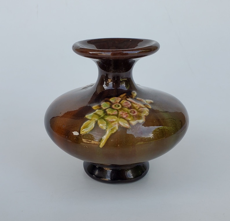 May include: A small, brown ceramic vase with a floral design. The vase has a wide base and a narrow neck. The floral design is in shades of green, yellow, and pink.