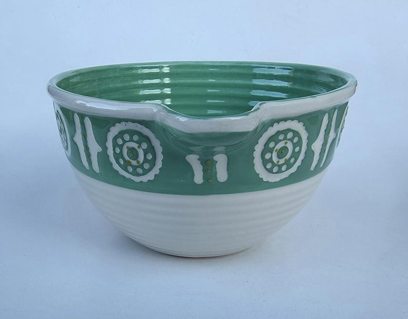 Peut inclure: Un saladier en c&eacute;ramique blanc et vert. Le bol pr&eacute;sente une base blanche avec des rainures horizontales et une partie sup&eacute;rieure verte. Des motifs circulaires et verticaux blancs d&eacute;corent la bande verte. Le bord est blanc et le bol a un bec verseur.