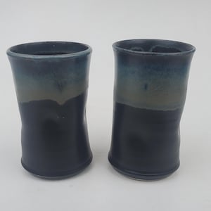 May include: Two handmade ceramic tumblers with a dark blue and gray gradient glaze. The cups have a slightly irregular shape and a smooth, glossy finish. The tumblers are suitable for cold or hot beverages.