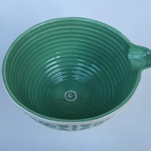 Peut inclure: Un saladier en c&eacute;ramique verte avec un bord blanc et une poign&eacute;e. Le bol a un int&eacute;rieur nervur&eacute; et un motif d&eacute;coratif au fond. Le bord ext&eacute;rieur pr&eacute;sente un motif r&eacute;p&eacute;titif. Le bol mesure environ 23 cm de diam&egrave;tre.