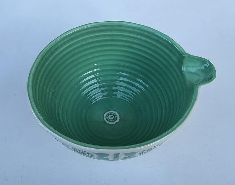 Peut inclure: Un saladier en c&eacute;ramique verte avec un bord blanc et une poign&eacute;e. Le bol a un int&eacute;rieur nervur&eacute; et un motif d&eacute;coratif au fond. Le bord ext&eacute;rieur pr&eacute;sente un motif r&eacute;p&eacute;titif. Le bol mesure environ 23 cm de diam&egrave;tre.