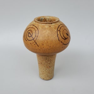 May include: A handmade ceramic vase with a mushroom-like shape. The vase is a speckled tan color with dark brown spiral designs on the rounded top. The vase has a long, cylindrical stem. The overall design is rustic and artistic.