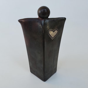 May include: A dark brown ceramic sculpture of a person with a heart on the chest. The heart is made of white ceramic and has a silver outline. The sculpture is inscribed with the words "The Call" on the base.