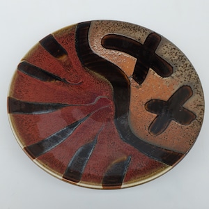 May include: A ceramic plate with a brown, red, and white glaze. The plate has a unique design with abstract shapes and lines.