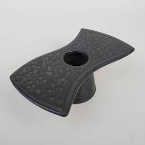 May include: A black ceramic vase with a textured surface and a round opening. The vase has a unique shape, resembling a butterfly with its wings spread.