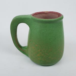 May include: A green ceramic mug with a rounded body and a large handle. The interior rim is a reddish-brown color. The mug has a textured surface and appears to be handmade. The mug is suitable for hot or cold beverages.
