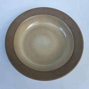 May include: A round ceramic bowl featuring a wide, dark brown rim and a lighter, speckled beige center. The bowl has a smooth, matte finish and a deep, concave basin, viewed from directly above against a plain, light-colored background.