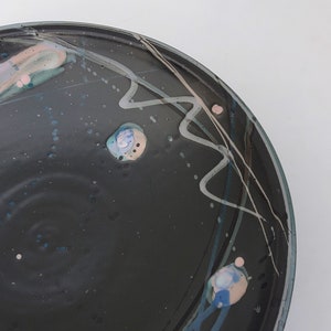 May include: A black ceramic bowl with a white, gray, and blue abstract design. The bowl has a speckled surface and a slightly raised rim.