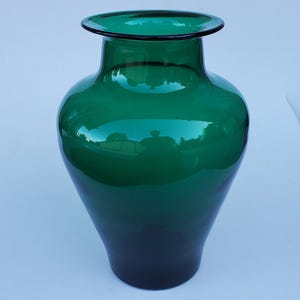 May include: A tall, emerald green glass vase with a wide, rounded body and a flared rim. The vase has a smooth, glossy finish and a dark green base. The vase is approximately 12 inches tall.
