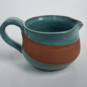 May include: A ceramic creamer pitcher featuring a glossy teal glaze on the interior and upper rim, contrasted by a wide, textured band of unglazed terracotta-colored clay around the center, complete with a curved handle and a small pouring spout.