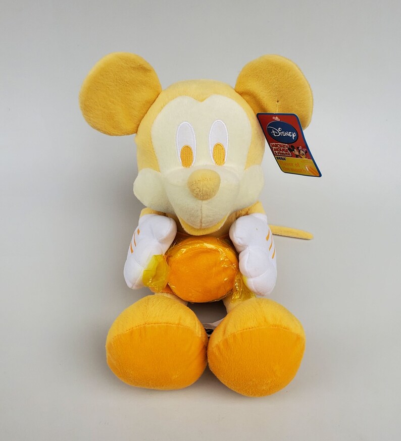 Puede incluir: Juguete de peluche amarillo de Mickey Mouse sosteniendo una pelota amarilla. El juguete tiene una cara blanca con ojos negros y una nariz negra. El juguete lleva una camisa blanca con rayas amarillas. El juguete tiene una etiqueta de Disney adherida a su oreja derecha.