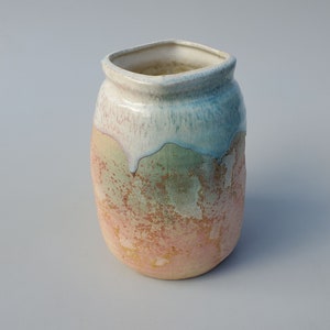 May include: A small, ceramic vase with a square opening and a rounded body. The vase has a white glaze with a blue and green drip effect, and a pink and orange speckled glaze on the bottom.
