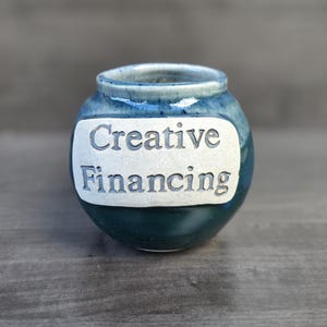 May include: A small, round ceramic jar with a blue and teal glaze. The jar features the words "Creative Financing" in a white, raised font on a white oval. The jar is sitting on a wooden surface.