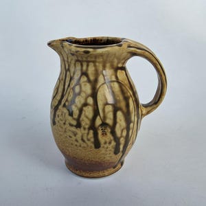 May include: A ceramic pitcher with a handle and spout. The pitcher has a beige base with dark brown drip glaze patterns. The rim and handle are a darker brown. The pitcher has a textured surface and a small design near the base.