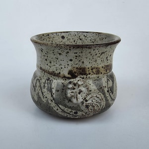 May include: A small, round ceramic cup with a speckled beige and brown finish. The upper portion features a flared rim, while the lower body is decorated with embossed circular floral motifs and dark gray abstract brushwork patterns.