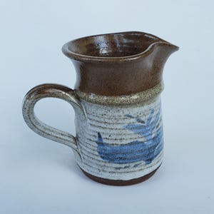May include: A handmade ceramic pitcher with a brown rim and handle. The body is white with blue abstract designs. The pitcher has a spout for easy pouring and is suitable for serving beverages. The pottery has a rustic, handcrafted appearance.