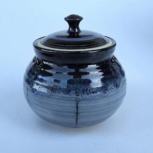 May include: A round ceramic sugar bowl with a lid, featuring a glossy dark navy blue glaze on the upper portion that transitions into a lighter, muted grey-blue tone on the lower body, characterized by subtle horizontal ridges and vertical lines.