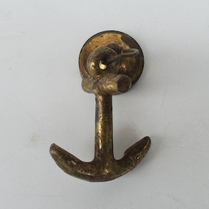 May include: A vintage brass anchor-shaped button with a circular base and a small loop. The anchor has a textured, aged appearance, with a central shaft and two curved arms. The button is likely for nautical-themed apparel or accessories.
