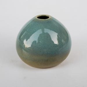 May include: A small, rounded ceramic vase with a teal and tan glaze. The vase has a narrow opening at the top and a smooth, glossy finish. The shape is reminiscent of a pebble or egg.