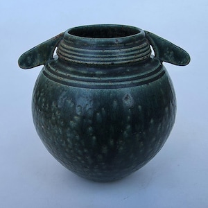 May include: A dark green ceramic vessel with a rounded body and two side handles. The surface features a mottled texture with lighter green spots and horizontal ridges encircling the neck area below the opening of the container.