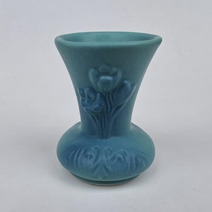 May include: A matte teal ceramic vase with a flared rim and a rounded base. The exterior features a raised relief design of a floral bouquet on the neck and a decorative scrolling pattern around the lower body of the vessel.