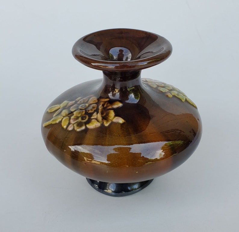 May include: A brown ceramic vase with a wide base and a narrow neck. The vase is decorated with raised floral designs in shades of green and yellow.