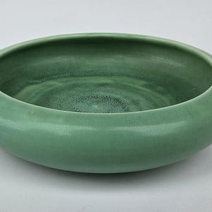 May include: A round, shallow, ceramic bowl in a muted green color. The bowl has a smooth, glossy finish and a wide, rounded rim. The interior of the bowl has a slightly textured appearance. This decorative bowl is suitable for serving or display.