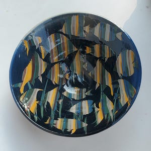 May include: A circular, dark blue ceramic bowl featuring a painted design of fish. The fish are decorated with vertical stripes in yellow, blue, green, and brown. The bowl's rim is a darker shade of blue.