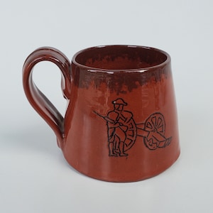 May include: A brown ceramic mug with a black silhouette of a person pushing a wheelbarrow. The mug has a handle and a rounded shape.