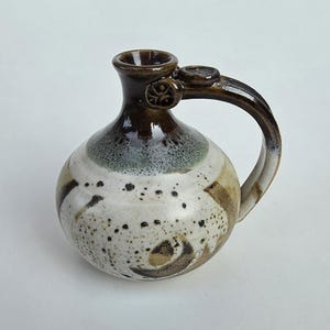 May include: A round ceramic pitcher with a narrow neck and a single handle. The body features a speckled cream and beige glaze with dark brown and green accents. The neck and handle are finished in a glossy dark brown glaze with a small circular detail.