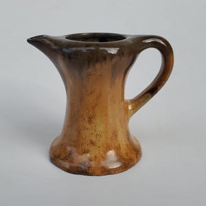 May include: A small, brown and yellow ceramic pitcher with a wide spout and a handle. The pitcher has a glazed finish and a rustic look.