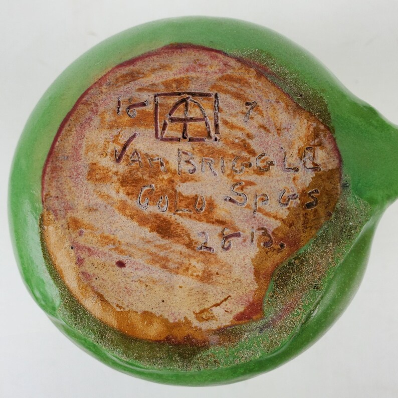May include: A green ceramic piece with a rounded shape and a handle. The bottom features text that reads "Van Briggle Gold Spgs 1613" and a square symbol. The rim has a reddish-brown color.