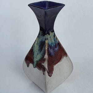 May include: A ceramic vase with a square top and a flared neck. The vase has a white base, transitioning to a reddish-brown and green midsection, and a dark blue top. The vase has a unique, geometric shape.