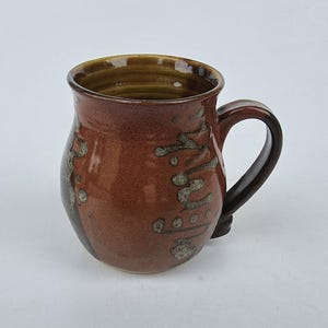 May include: A handmade ceramic mug with a brown glaze and decorative speckled accents. The mug has a rounded body, a dark brown handle, and a capacity of approximately 12 ounces. The interior of the mug is a darker brown.