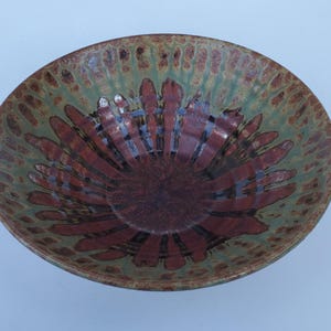 May include: A handmade ceramic bowl with a unique design. The bowl features a dark brown center with radiating patterns in a reddish-brown hue. The outer rim has a speckled green and brown pattern.