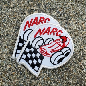 May include: Two white iron-on patches with the word "NARC" in red lettering. One patch features a cartoon illustration of a race car driver in a red racing suit driving a white race car with a checkered flag.