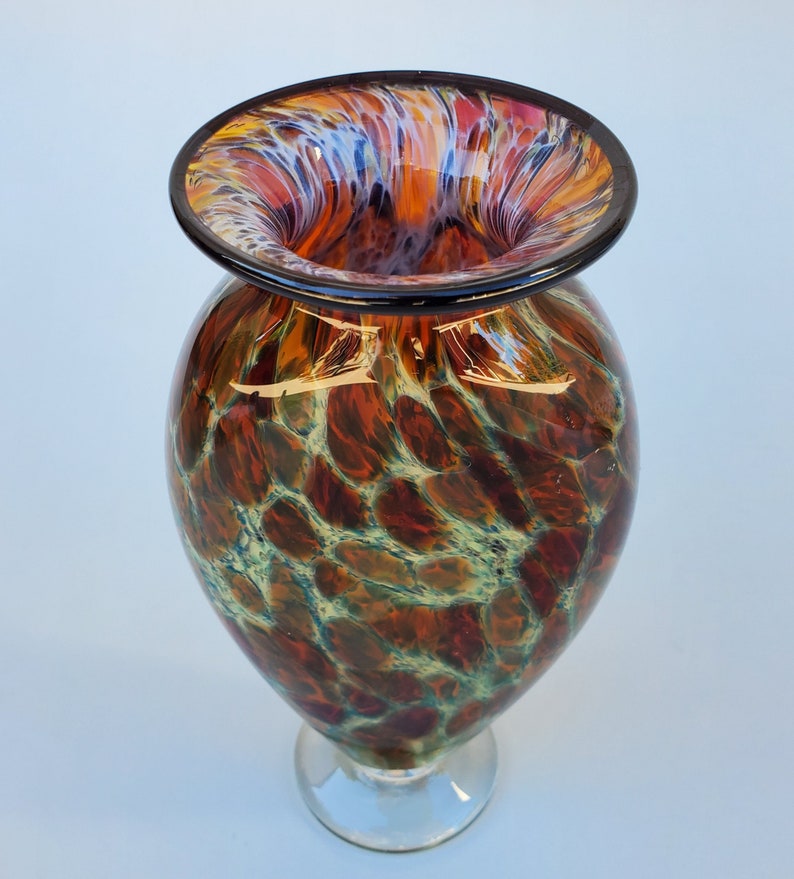 May include: A tall, brown and green glass vase with a mottled, textured surface. The vase has a wide, flared opening with a black rim and a white, blue, and red swirl pattern.