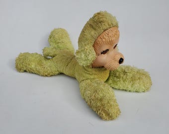 May include: A green plush toy dog with a brown face and floppy ears. The dog is lying on its belly with its head turned to the side.