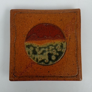 May include: Square ceramic coaster with a speckled brown background and a circular design. The circle features a two-tone landscape with a red upper half and a green and black lower half. The coaster has a raised edge.