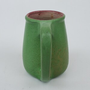 May include: A green ceramic mug with a handle. The mug has a textured surface and a slightly tapered shape. The interior rim is a reddish-brown color. The mug is likely handmade and suitable for hot or cold beverages.