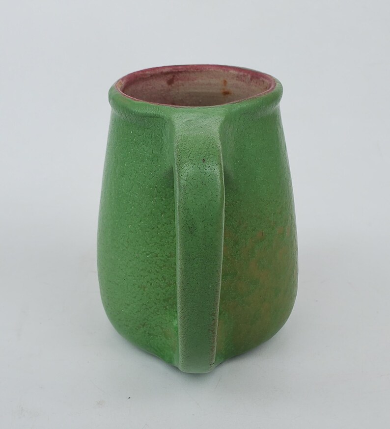 May include: A green ceramic mug with a handle. The mug has a textured surface and a slightly tapered shape. The interior rim is a reddish-brown color. The mug is likely handmade and suitable for hot or cold beverages.
