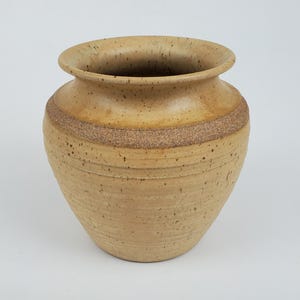May include: A handcrafted, beige ceramic vase with a wide opening and a textured, speckled surface. A band of darker, rough texture encircles the middle. The vase has a rounded body and a slightly flared rim, suitable for home decor.