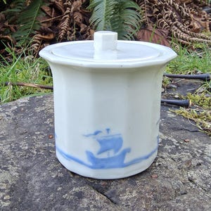 May include: A white ceramic jar with a lid and a blue sailboat design. The jar has a cylindrical shape with a slightly flared rim and a small, square handle on the lid. The sailboat design is repeated around the base of the jar.