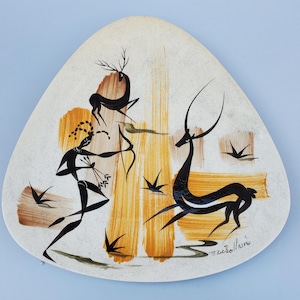 May include: A triangular ceramic wall plate featuring a stylised black and white design of a hunter with a bow and arrow, two deer, and stylised birds. The plate has a white background with brown and yellow accents. The plate is signed "Macerolline".