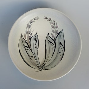 May include: A white ceramic plate with a stylized floral design in shades of green and gray. The design features a cluster of leaves and flower buds.